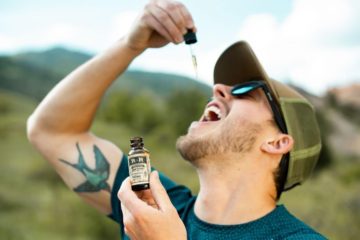 Guy in his 20's or early 30's with tattoo of bird on right inner bicep wearing a baseball cap and sunglasses holding bottle of cbd tincture and using eyedropper to apply under tongue.