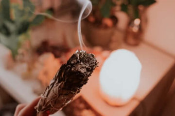 Women's hand holding burning sage with desk in the background with plants and a light on it.