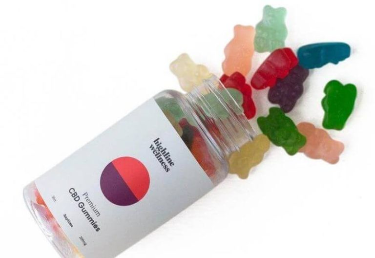 Clear bottle of Highline Wellness CBD gummy bears laying on its side with assorted gummy bears on white background.