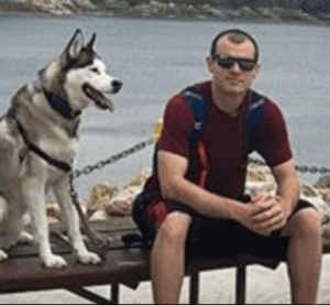 Josh Murdoch the founder of Certified CBD in a maroon tshirt black shorts a blue camelback and sunglasses on sitting on a bench next to his Siberian husky named Ace with a lake in the background.