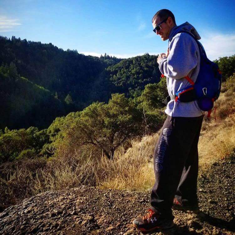 Josh Murdoch the founder of Certified CBD wearing a blue camelbak grey sweatshirt black sweat pants and black and orange under armor sneakers standing on a hiking trail looking down the side of a mountain.