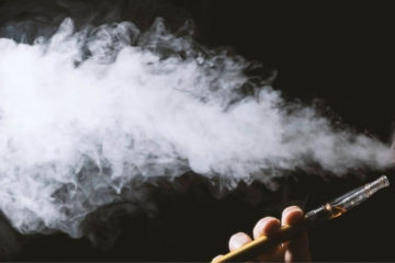 Guy holding a CBD vape pen in his hand blowing a cloud of vape smoke .