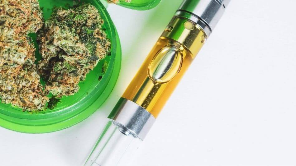 Cannabis vape pen laying next to a green plastic grinder full of hemp flower.