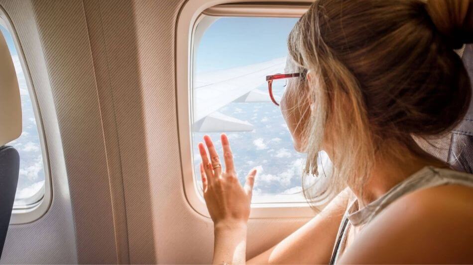 Women in her twenties with dirty blonde hair sitting in the window seat on an airplane with one hand touching the window while she stairs out at the beautiful view.
