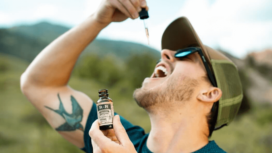 Guy in his 20's or early 30's with tattoo of bird on right inner bicep wearing a baseball cap and sunglasses holding bottle of CBD tincture and using eyedropper to apply under tongue.