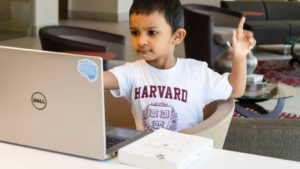 Young kid in a white Harvard shirt with red writing sitting in front of a silver laptop holding one of his hands up to ask a question.