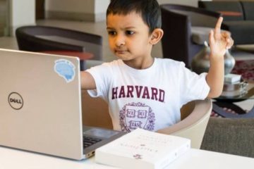 Young kid in a white Harvard shirt with red writing sitting in front of a silver laptop holding one of his hands up to ask a question.