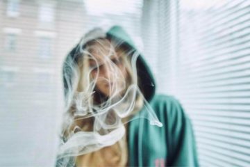 Blonde women wearing a green hoodie and the camera focused on smoke floating in front of her.