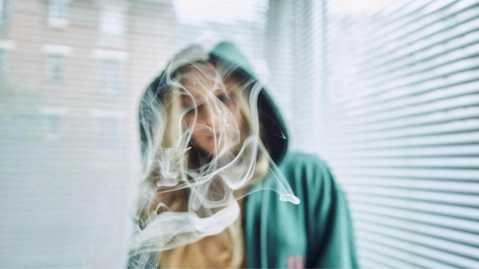 Blonde women wearing a green hoodie and the camera focused on smoke floating in front of her.