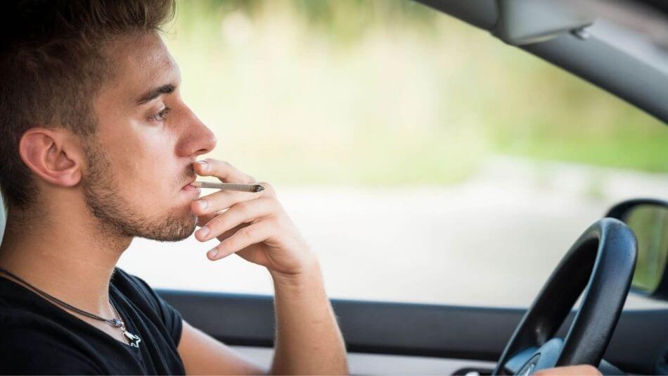 Young guy in his twenties wearing a black tshirt driving a car and smoking a joint.