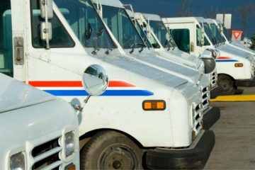 A line of white USPS trucks lined up parked in a parking lot.