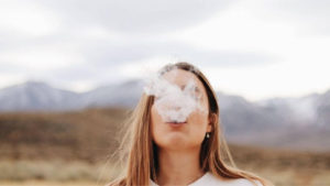 Strawberry blonde women blowing out a cloud of smoke a mountain range in the background.