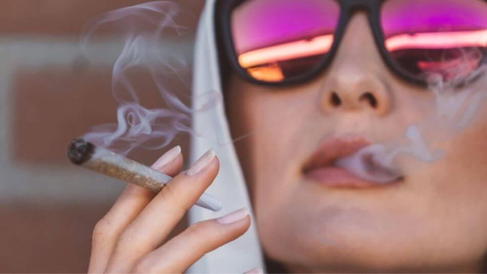 Women wearing sunglasses with pink lenses smoking a joint blowing out smoke.