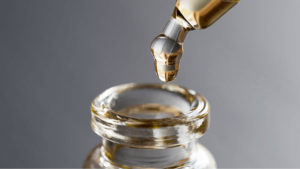 Zoomed in view of CBD oil dripping from glass eyedropper into glass bottle with grey background.