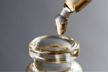 Zoomed in view of CBD oil dripping from glass eyedropper into glass bottle with grey background.