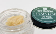 Plain Jane og kush CBD crumble in the jar with the lid behind and to the right.