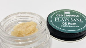 Plain Jane og kush CBD crumble in the jar with the lid behind and to the right.