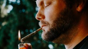 Man with a scruffy beard lighting a joint that's hanging from his lips.