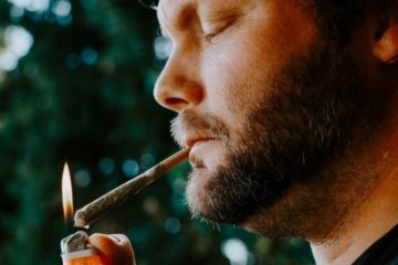 Man with a scruffy beard lighting a joint that's hanging from his lips.