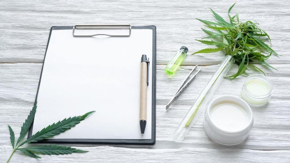 Clipboard with a blank sheet of paper and a pen sitting on top next to a test tube with cannabis.