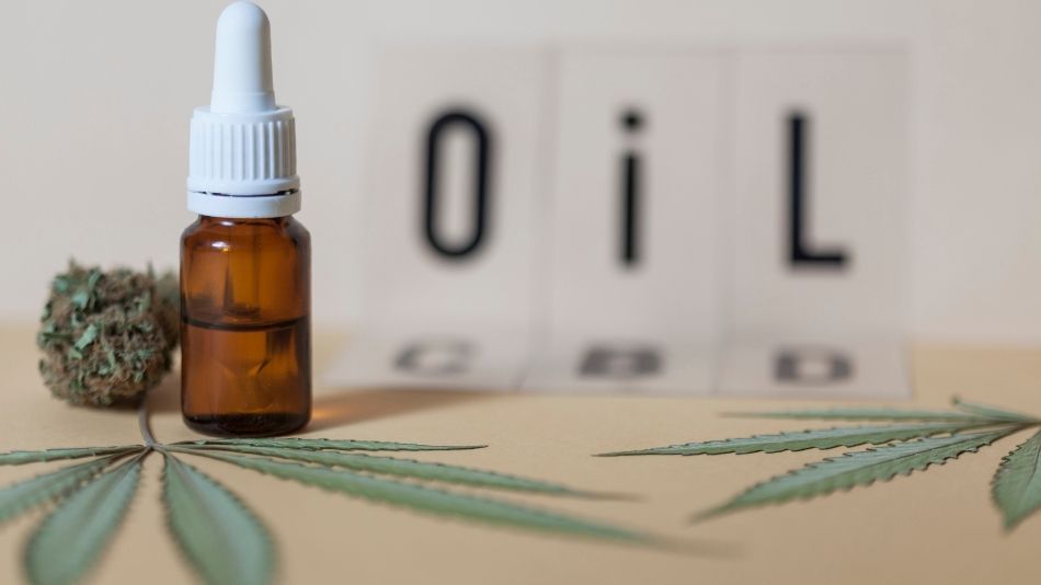 Bottle of CBD oil standing on the table with two cannabis leafs laying on the table and the words CBD oil behind.