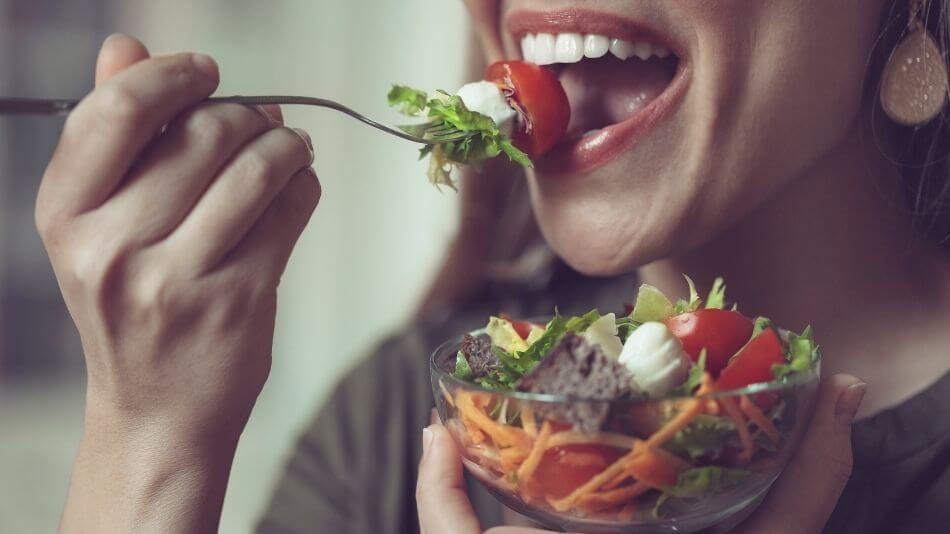 Women holding a bowl of salad in one hand and eating a bite full on a fork with the other.
